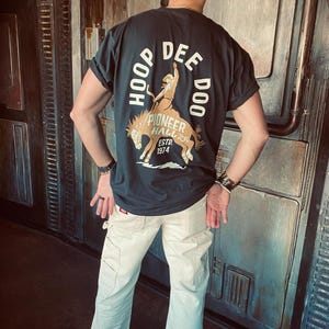 Könnte beinhalten: Eine Person trägt ein schwarzes T-Shirt mit einem weißen Aufdruck eines Cowboys, der ein Pferd reitet. Der Aufdruck lautet "HOOP DEE DOO" und "PIONEER HALL ESTD. 1974". Die Person trägt eine weiße Hose.