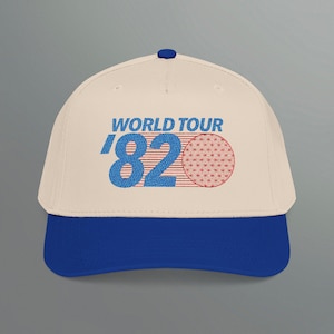 May include: Cream-colored baseball cap with a blue brim and button. The cap features the text "WORLD TOUR '82" in blue and red, along with a red geometric design. A stylish accessory for casual wear.