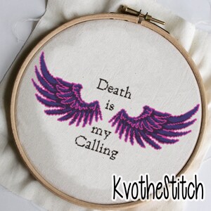May include: Embroidered hoop art with purple and pink angel wings and the text "Death is my Calling". The hoop is on a white fabric background.