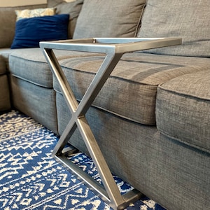 May include: A silver metal table with an X-shaped base. The table is placed on a gray couch with a blue pillow. The table is sitting on a blue and white patterned rug.