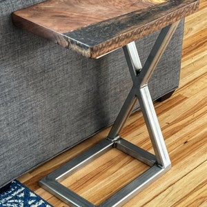 May include: A wooden side table with a live edge top and a dark epoxy resin inlay. The table has a metal base with an X-shaped design.