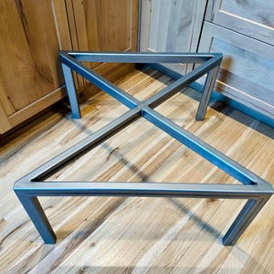 May include: A pair of unfinished metal table legs. The legs are constructed from square tubing and feature a geometric design with an X-shaped cross brace. The legs are designed to be used as a base for a table or other furniture.