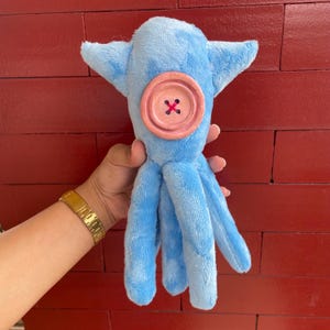 May include: A blue plush octopus with a pink button for an eye. The octopus has six tentacles.