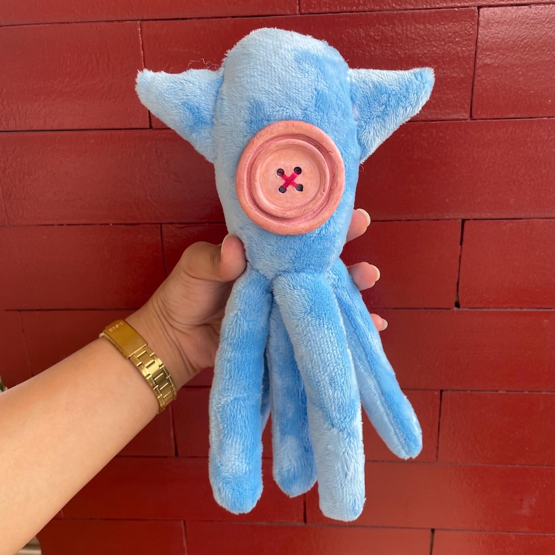 May include: A blue plush octopus with a pink button for an eye. The octopus has six tentacles.