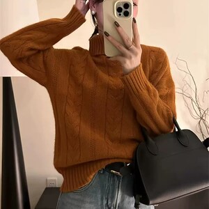 Könnte beinhalten: Eine Frau trägt einen rostfarbenen Zopfmuster-Rollkragenpullover und eine hellblaue Jeans. Eine schwarze Handtasche mit Henkel und ein schwarzer Gürtel mit goldener Schnalle sind ebenfalls zu sehen. Die Frau hält ein Telefon vor ihr Gesicht.