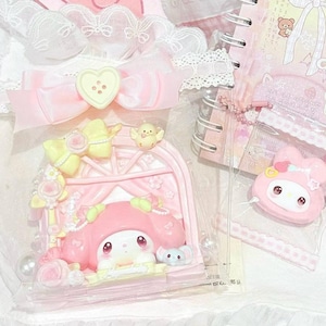 Op de afbeelding: Een verzameling roze en witte briefpapierartikelen, waaronder een notitieboek met "Hello Kitty" op de omslag, een hartvormig haaraccessoire met "My Melody"-tekst en een decoratief My Melody-beeldje. De items zijn gerangschikt met kant en linten.