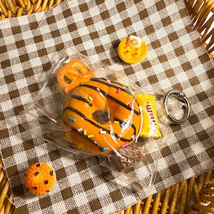 May include: An orange donut with chocolate drizzle and sprinkles, a pretzel, a cookie, and a small candy bar are in a clear plastic bag. A yellow tart with a cat on top and a silver ring are also present. The items are arranged on a brown and white checkered cloth inside a wicker basket.