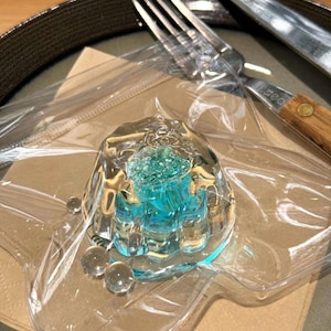 May include: A clear plastic container with a blue jelly dessert inside. The jelly has a white and green design. The container is on a brown paper napkin.