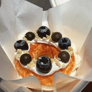 May include: A ring-shaped donut with a golden-brown crust, decorated with white cream, fresh blueberries, and chocolate-covered sweets. The donut is sprinkled with chopped almonds and served in a white paper container.