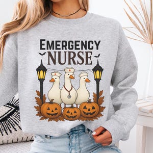 Może przedstawiać: Popielata szara bluza z napisem "EMERGENCY NURSE" nad grafiką przedstawiającą trzy gęsi w czapkach pielęgniarskich i stetoskopach, z dyniami i lampionami. Słowo "Ash" jest wydrukowane w lewym dolnym rogu.