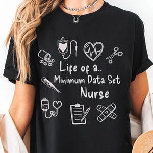 Può includere: T-shirt nero con illustrazioni grafiche e testo bianchi. Il testo recita "Life of a.. Minimum Data Set Nurse". La maglietta presenta icone a tema medico come un monitor cardiaco, forbici e un termometro.