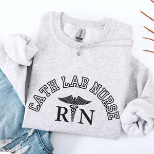 Könnte beinhalten: Ein graues Sweatshirt mit dem Text "Cath Lab Nurse RN" in schwarzen Buchstaben mit einem Caduceus-Symbol. Das Sweatshirt liegt auf einer weißen Oberfläche mit einer blauen Jeans.