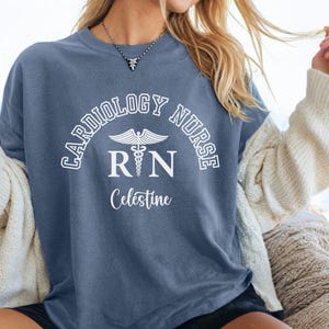 May include: A blue t-shirt with white text that reads "Cardiology Nurse RN Celestine". The text is in an arc shape with a caduceus symbol in the center.