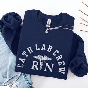 Könnte beinhalten: Marineblaues Sweatshirt mit dem weißen Text "CATH LAB CREW RN" und einem Caduceus-Symbol. Eine silberne Halskette mit Anhänger liegt auf dem Sweatshirt. Das Sweatshirt liegt gefaltet auf einer Jeans.