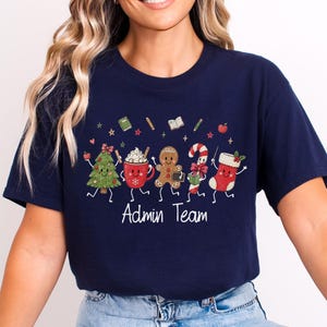 May include: A navy blue t-shirt featuring a graphic of five animated Christmas characters including a tree, a mug, a gingerbread person, a candy cane, and a stocking, with the text "Admin Team" printed in white below the festive illustration.