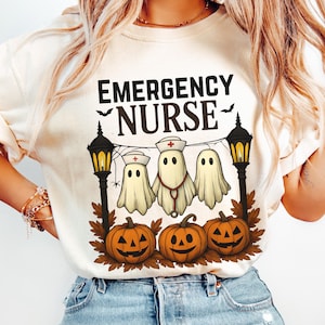 May include: Ivory-colored t-shirt with a Halloween-themed graphic. The design features the text "EMERGENCY NURSE" above three cartoon ghosts wearing nurse hats and a stethoscope, with jack-o'-lanterns below. Two black street lamps flank the design.