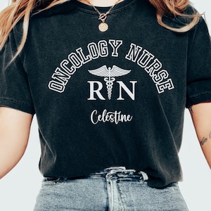 May include: Black t-shirt with white text that reads "Oncology Nurse RN Celestine". The text is in an arc shape with a caduceus symbol in the centre. The shirt is being worn by a person with blue jeans.