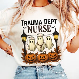 Może przedstawiać: Kremowa koszulka z napisem "TRAUMA DEPT NURSE" nad grafiką trzech postaci duchów przebranych za pielęgniarki, z dyniami. Projekt zawiera czarne latarnie i nietoperze, idealne na Halloween.