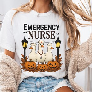 Może przedstawiać: Biała koszulka z napisem "EMERGENCY NURSE" nad grafiką przedstawiającą trzy kaczki w czapkach pielęgniarskich, ze stetoskopami i dyniami. Projekt zawiera czarne latarnie i jesienne liście, idealny na Halloween.