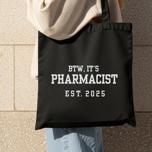 Peut inclure: Sac fourre-tout noir avec le texte "BTW. IT'S PHARMACIST EST. 2025" en lettres blanches. Le sac a des poignées noires et est porté par une personne.