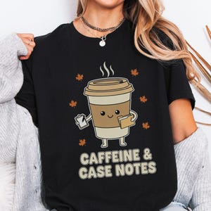 Könnte beinhalten: Ein schwarzes T-Shirt mit einer Grafik einer lächelnden Kaffeetasse, die ein Klemmbrett und einen Aktenordner hält, umgeben von kleinen orangefarbenen Ahornblättern. Unter der Grafik steht der Text CAFFEINE & CASE NOTES in heller Schrift.