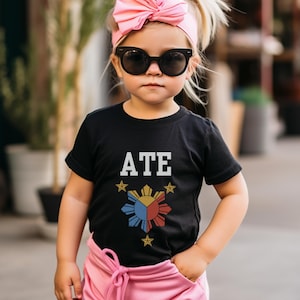 May include: A black t-shirt with a white "ATE" graphic and a stylized image of the Philippine flag. The shirt is being worn by a young child with pink hair and a pink headband.