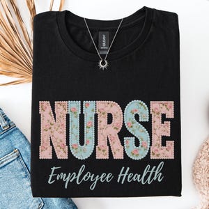 May include: Black t-shirt with the word "NURSE" in large, floral-patterned letters. The word "NURSE" is in pink and blue floral print. Below the word "NURSE" is the text "Employee Health" in a script font. A silver necklace is visible.