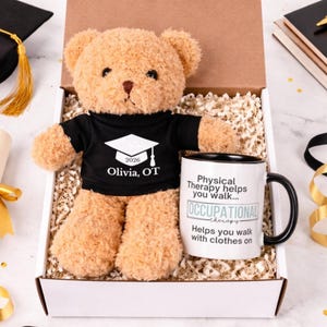 May include: A teddy bear wearing a black t-shirt with a graduation cap graphic and the text "2026 Olivia, OT" sits in a gift box with a mug. The mug has the text "Physical Therapy helps you walk... OCCUPATIONAL therapy Helps you walk with clothes on."