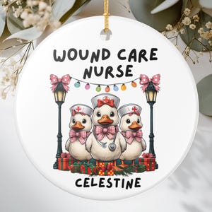 May include: White ceramic Christmas ornament featuring three cartoon ducks dressed as nurses, with the text "WOUND CARE NURSE" and "CELESTINE". The ducks wear nurse hats and pink bow ties, with a string of lights above them.