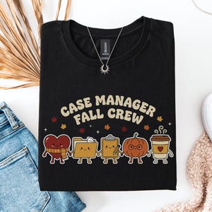 Może przedstawiać: Czarny t-shirt z napisem "CASE MANAGER FALL CREW" w beżowym kolorze. Pod tekstem znajduje się pięć postaci z kreskówek: serce z szalikiem, teczka na dokumenty, podkładka z ołówkiem, dynia w okularach oraz kubek z kawą.