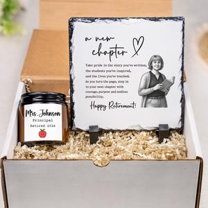 Op de afbeelding: Een cadeaupakket met een kaars en een leistenen plaat. De kaars heeft het opschrift "Mrs. Johnson, Principal, Retired 2026" en een rode appel illustratie. De plaat toont de tekst "a new chapter" en "Happy Retirement!"