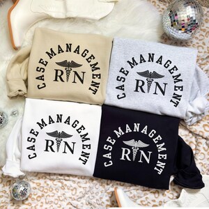 May include: Four sweatshirts in beige, gray, white, and black, each with the text "CASE MANAGEMENT RN" in a semi-circular design. The RN is centered below a caduceus symbol. The sweatshirts are folded on a patterned surface.