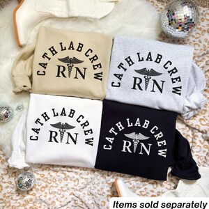 Könnte beinhalten: Vier Crewneck-Sweatshirts in Beige, Grau, Weiß und Schwarz. Jedes Sweatshirt hat den Text "CATH LAB CREW RN" in einem Bogen mit einem medizinischen Symbol in der Mitte aufgedruckt. Die Sweatshirts werden auf einer gemusterten Oberfläche präsentiert.