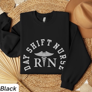 Könnte beinhalten: Schwarzes Sweatshirt mit dem Text "DAY SHIFT NURSE RN" in einem Halbkreis-Design. Das Design enthält ein Caduceus-Symbol. Das Wort "Black" befindet sich in der unteren linken Ecke des Bildes.
