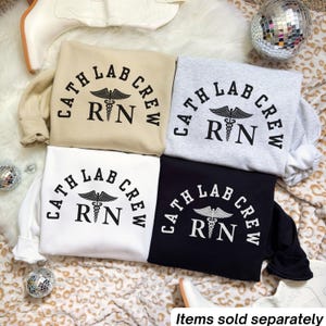 Könnte beinhalten: Vier Sweatshirts in Beige, Grau, Weiß und Schwarz, jeweils mit dem Text "CATH LAB CREW RN" in einem kreisförmigen Design mit einem medizinischen Symbol. Die Sweatshirts werden auf einer gemusterten Oberfläche präsentiert.