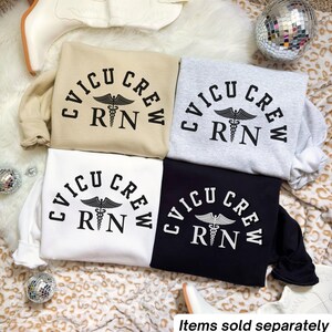 Könnte beinhalten: Vier Sweatshirts in Beige, Grau, Weiß und Schwarz, jeweils mit dem Text "CVICU CREW RN" über einem medizinischen Caduceus-Symbol. Die Sweatshirts werden auf einer gemusterten Oberfläche präsentiert.