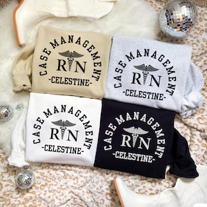 Puede incluir: Cuatro sudaderas en beige, gris, blanco y negro, cada una con el texto "CASE MANAGEMENT" en semicírculo, las letras "RN" con un símbolo de caduceo y "-CELESTINE-" debajo. Las sudaderas se muestran sobre una superficie estampada.