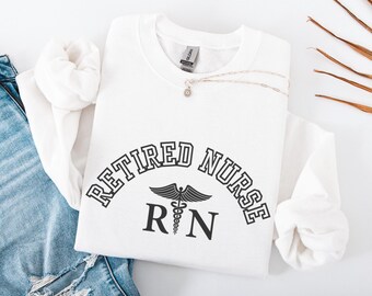 Puede incluir: Una sudadera blanca con la inscripción negra "RETIRED NURSE" en arco sobre un símbolo de caduceo y las letras "RN". Un collar plateado con un pequeño colgante descansa sobre la sudadera. La palabra "White" está en la esquina inferior izquierda.
