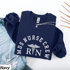 Può includere: Felpa blu navy con la scritta bianca "MDS Nurse Crew RN" e un simbolo di caduceo. Una collana d'argento con un ciondolo è drappeggiata sulla felpa. La felpa è su una superficie bianca con un paio di jeans.