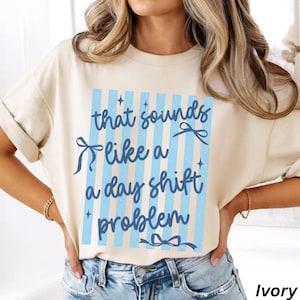 May include: An ivory short-sleeved t-shirt featuring a central graphic with vertical light blue stripes. The text "that sounds like a day shift problem" is printed in dark blue cursive, accented by small blue bows and star-shaped sparkles.
