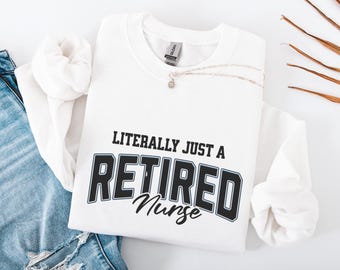 Puede incluir: Sudadera blanca con el texto "LITERALLY JUST A RETIRED Nurse" en letras negras y grises. Un collar plateado con un pequeño colgante descansa sobre la camisa. La sudadera está doblada sobre unos vaqueros azules.