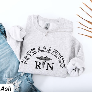 Könnte beinhalten: Hellgraues Sweatshirt mit dem Text "CATH LAB NURSE" in einem Bogen, darunter ein RN-Symbol. Das Sweatshirt liegt gefaltet auf einer blauen Jeans. Eine silberne Halskette mit einem kleinen Anhänger liegt auf dem Sweatshirt.