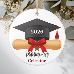 Può includere: Ornamento di laurea rotondo in ceramica bianca. Presenta un cappello nero con nappina rossa, un diploma arrotolato e l'anno 2026. L'ornamento ha anche un fiocco rosso e le parole "Phlebotomist Celestine."