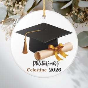 Può includere: Ornamento di laurea in ceramica bianca con un cappello di laurea nero con nappina dorata e un diploma arrotolato legato con un nastro dorato. Il testo recita "Phlebotomist Celestine 2026".