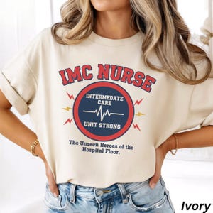 Może przedstawiać: Kremowa koszulka z napisem "IMC NURSE" w kolorze czerwonym i niebieskim. Projekt zawiera okrągłą grafikę z napisem "Intermediate Care" i linią bicia serca. Obecne są również teksty "Unit Strong" i "The Unseen Heroes of the Hospital Floor".
