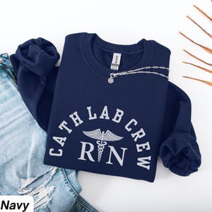 Könnte beinhalten: Marineblaues Sweatshirt mit weißem Text "CATH LAB CREW RN" und einem Caduceus-Symbol. Das Sweatshirt liegt gefaltet auf einer Jeans. Eine silberne Halskette mit Anhänger liegt auf dem Sweatshirt.