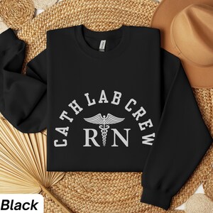 Könnte beinhalten: Schwarzes Sweatshirt mit Rundhalsausschnitt und dem Text "CATH LAB CREW" in einem Halbkreis über dem medizinischen Symbol und den Buchstaben "RN" in Weiß. Das Wort "Black" befindet sich unten links.