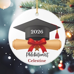 Può includere: Ornamento natalizio in ceramica bianca con un cappello di laurea con l'anno 2026, un diploma e un fiocco rosso. Il testo recita "Phlebotomist Celestine". Un filo d'oro è attaccato per appenderlo.