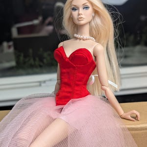 May include: A fashion doll with long blonde hair, blue eyes, and a pearl necklace. The doll wears a red velvet bodice and a layered pink tulle skirt. The doll is posed with arms at its sides.
