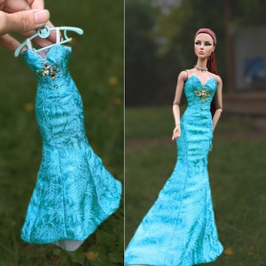 Puede incluir: Un vestido de sirena azul turquesa con un adorno de cuentas en la cintura. El vestido está en una percha y una muñeca lleva el mismo vestido.